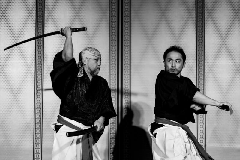 Two men in traditional Japanese attire are performing a dramatic sword scene; one holds a raised katana as if to strike, while the other appears ready to defend himself, both showing intense expressions.