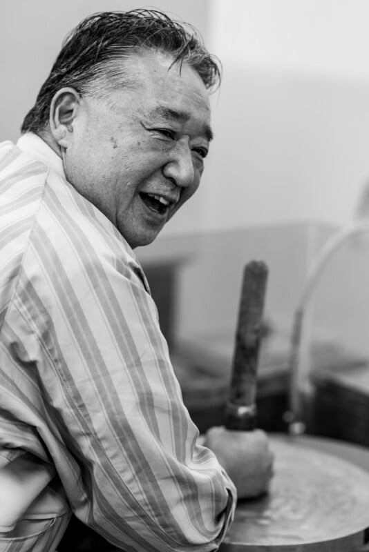 A man in a striped shirt smiles while working, using a wooden tool to shape or mix something in a large round container. The image is black and white, capturing a candid, joyful moment.