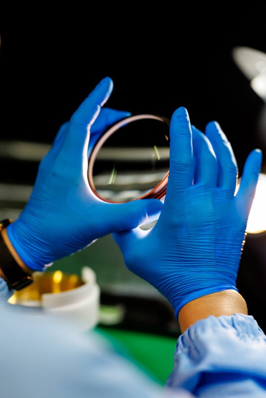 Person wearing blue gloves and a lab coat holds a transparent circular object, possibly a petri dish or lens, with both hands in a laboratory setting.