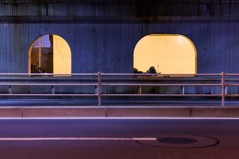 A person lies down inside a lit archway beneath an overpass at night, with another empty archway nearby. Metal railings and an empty street are visible in the foreground.