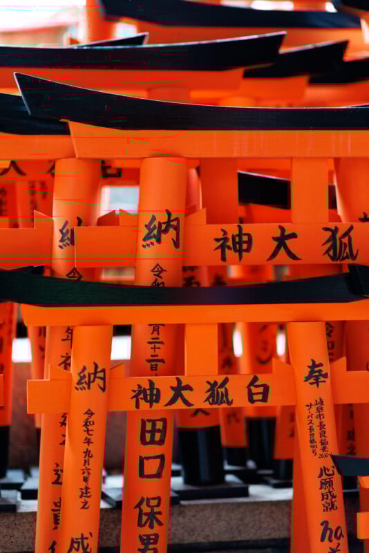A close-up of multiple small, bright orange torii gates with black Japanese inscriptions, stacked together at a shrine. The gates feature bold black tops and traditional calligraphy.