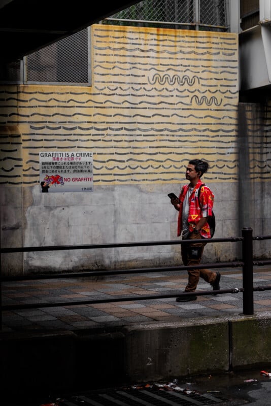 A man wearing a red and yellow shirt walks on a wet sidewalk, looking at his phone. Behind him is a wall with a “GRAFFITI IS A CRIME” sign and patterned yellow lines.