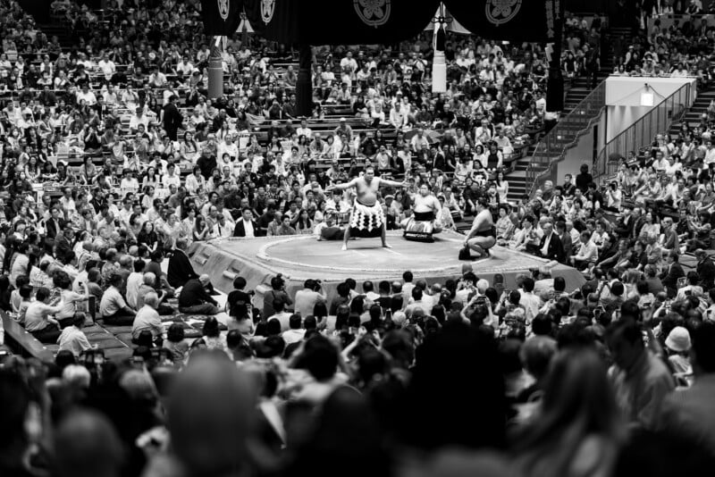 A large crowd watches a sumo wrestling match in an arena. Two sumo wrestlers face each other in the ring, with a referee and assistants present, all surrounded by spectators. The image is in black and white.