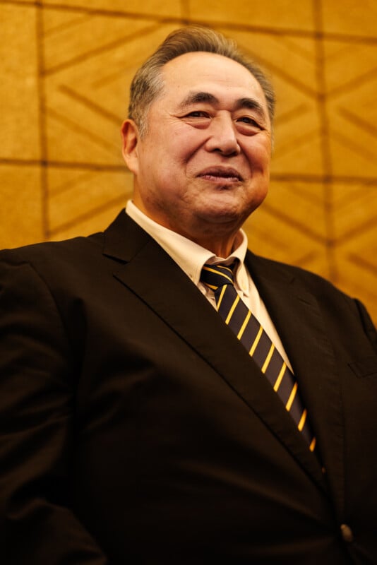 A man in a dark suit, light shirt, and striped tie stands in front of a textured gold background, smiling slightly and looking confidently ahead.