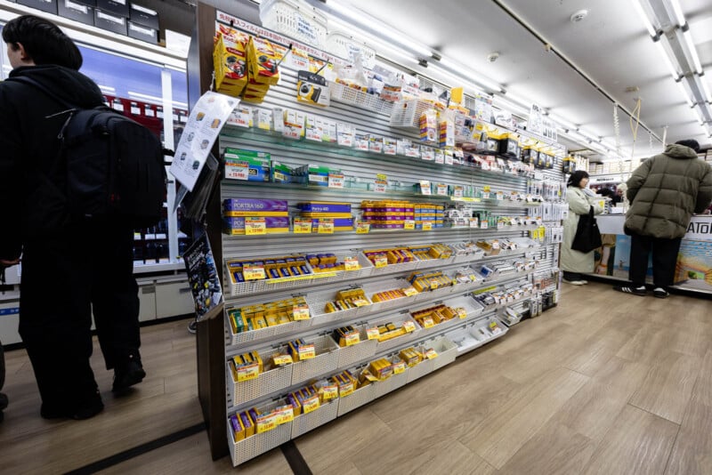 A well-lit pharmacy aisle with neatly organized shelves displaying various medicines and health products; several customers are browsing and checking out at the counter.