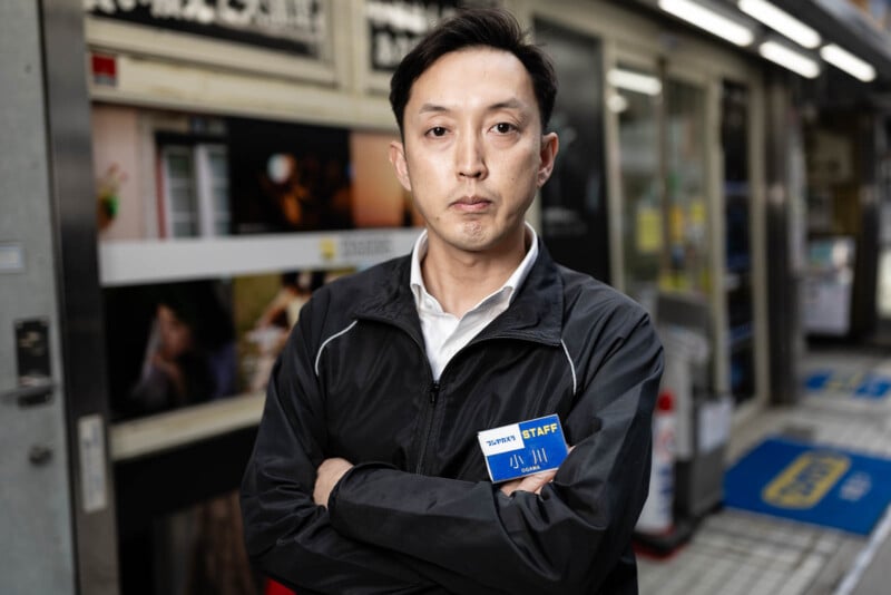 A man wearing a black jacket and a staff name badge stands with arms crossed in front of a store entrance, looking directly at the camera. The background shows store signs and part of a tiled walkway.