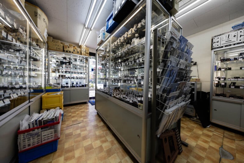 A brightly lit store interior with glass display cases filled with electronic components and accessories, shelves lined with small boxes, and plastic bins containing various items on the floor.