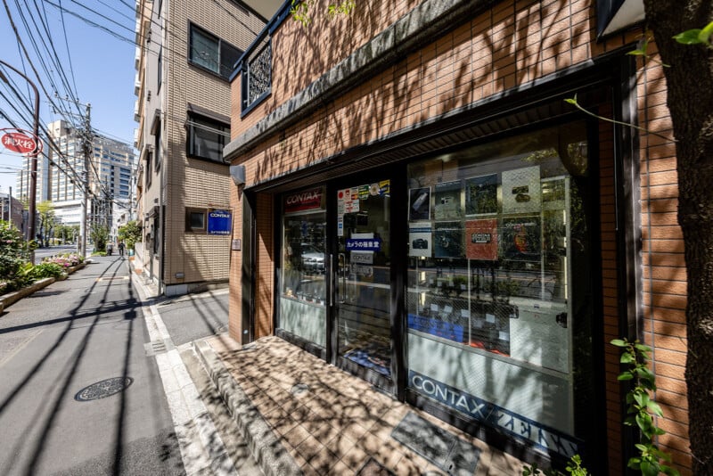 A small camera shop with glass doors and display windows sits on a quiet urban street lined with buildings and power lines. Signs and camera posters are visible in the windows. Trees cast shadows on the sidewalk.