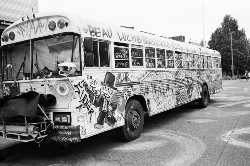A large bus covered in graffiti art and tags is parked on a street. The designs include bold text, abstract patterns, and a black-and-white cartoon figure in a hat painted near the front wheel.