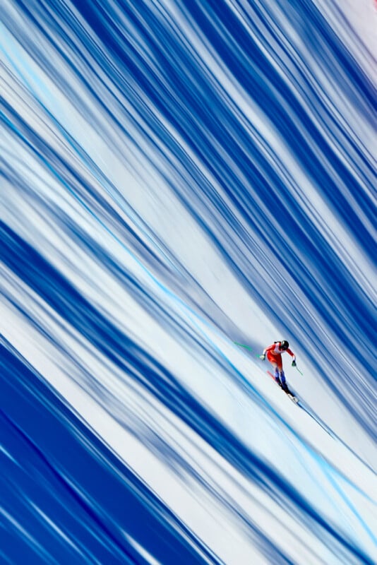 A skier in a red suit races downhill on a snow-covered slope, surrounded by blue and white streaks of shadow and light, creating a dynamic, abstract background.