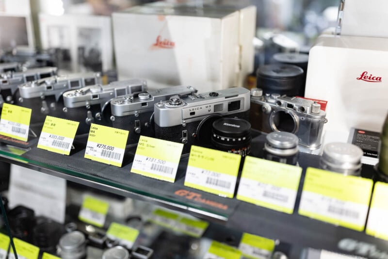 A display case with vintage Leica cameras, lenses, and accessories. The items have bright yellow price tags, and Leica branding is visible on some boxes and camera bodies in the background.