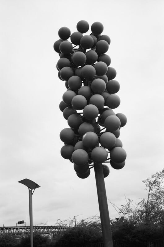 A tall outdoor sculpture composed of clustered spherical shapes on a vertical pole, set against a cloudy sky with trees and part of a bridge in the background.