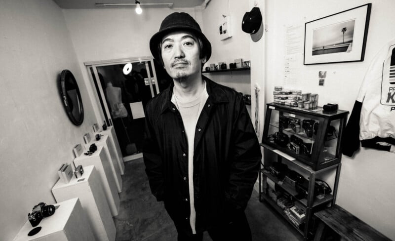 A man wearing a hat and jacket stands in a small, stylish room with vintage cameras, photos, and shelves in the background. The image is in black and white, giving it a classic, artistic feel.