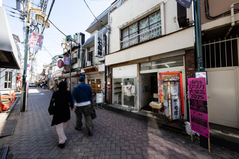 Two people walk down a narrow street lined with shops in Japan. Signs with Japanese text hang above storefronts, and a bright pink poster is attached to a pole on the right side of the street.