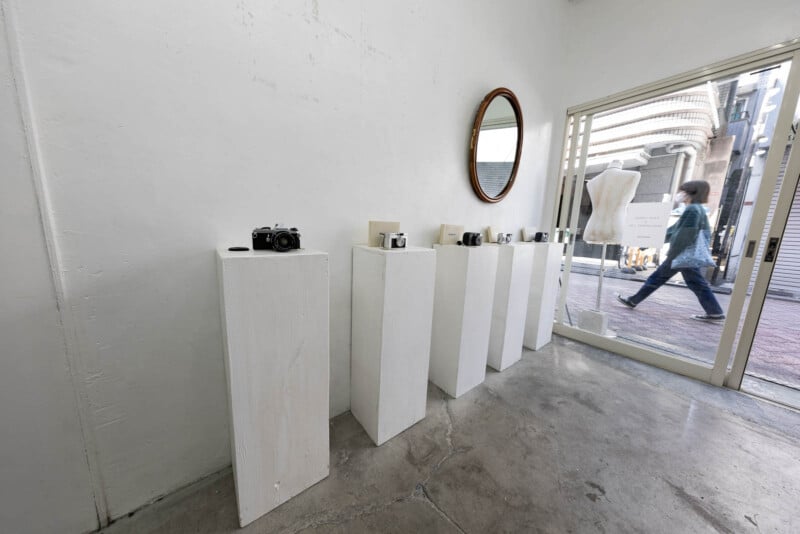 A row of vintage cameras is displayed on white pedestals in a minimalist gallery with white walls, a round mirror, and a mannequin near a glass door; a person walks past outside on the street.