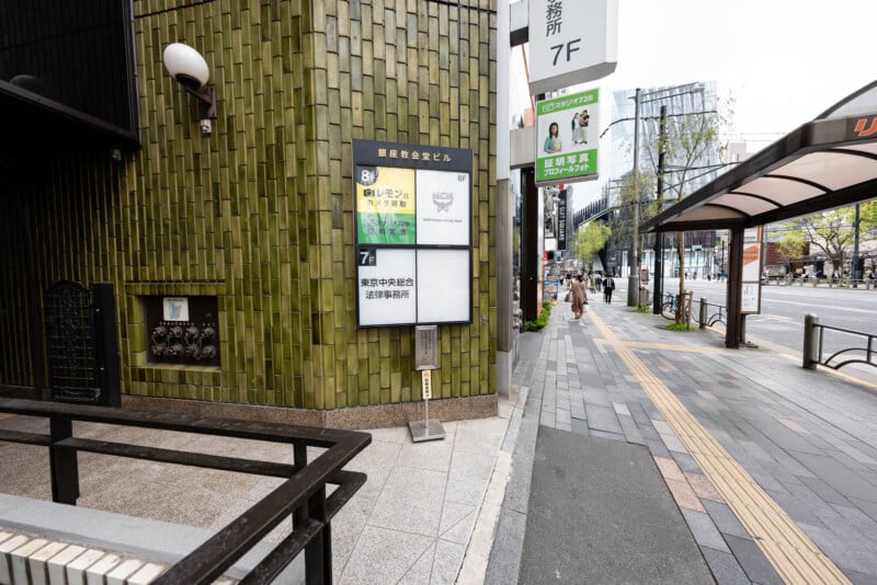 A city sidewalk in Japan with tiled green building, a large white and yellow signboard, a bus stop shelter, and trees lining the street on a cloudy day.