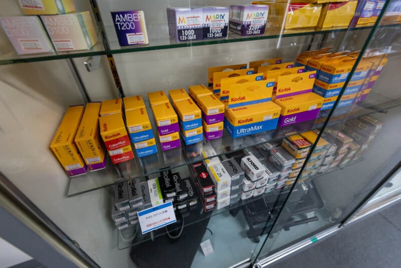 Boxes of Kodak and other brands of photographic film are displayed on glass shelves in a store, with yellow, purple, and white packaging visible. A label on one shelf reads "FILM FOR SALE IN BOX, EXP. VARIOUS.