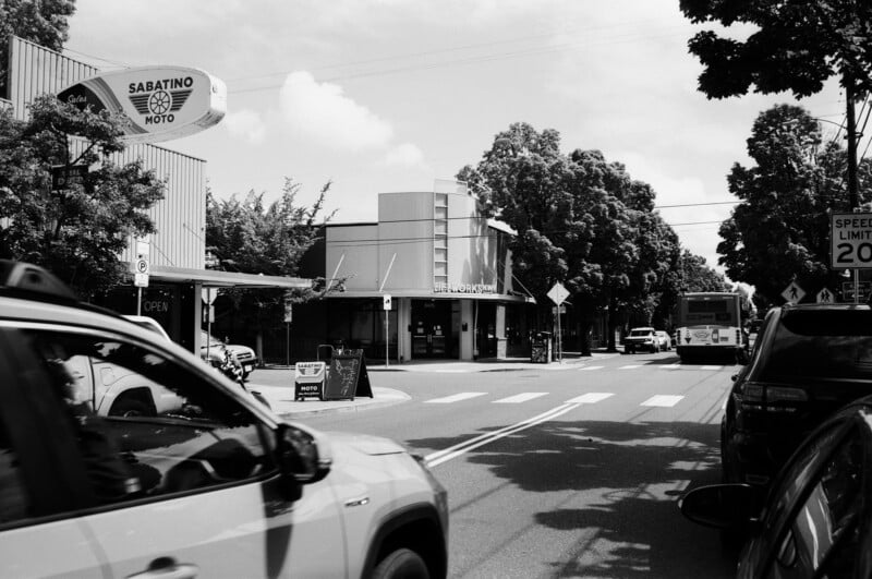 A black-and-white photo of a street with cars, a bus, and a pedestrian crossing. There are trees, shops, and a "Sabatino Moto" sign on the left, and a "Speed Limit 20" sign on the right.
