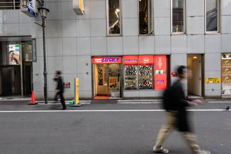 A street view of a "Lucky Camera" store with cameras displayed in the window. Two blurred pedestrians walk past, and a streetlamp and traffic cone are visible on the left side of the image.