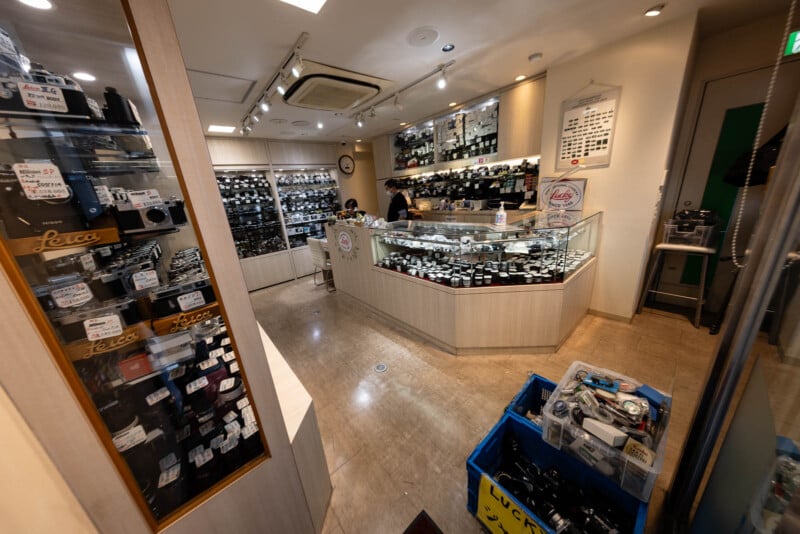 A camera store interior with glass display cases filled with cameras and lenses, a shopkeeper behind the counter, shelves of camera equipment, and a blue bin of used cameras on the floor in the foreground.