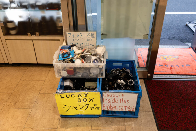 Two plastic boxes near a shop entrance; one labeled “LUCKY BOX” contains camera accessories, the other, labeled “Caution!! Inside this Broken camera,” holds broken camera parts. Signs with Japanese and English text are visible.