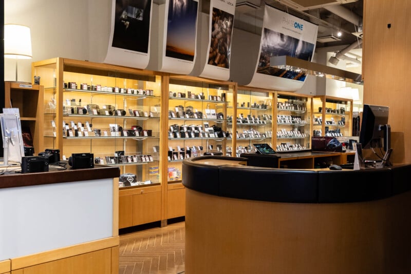 A well-lit camera store interior featuring glass display cases filled with cameras and accessories, with a wooden counter and computer visible in the foreground.