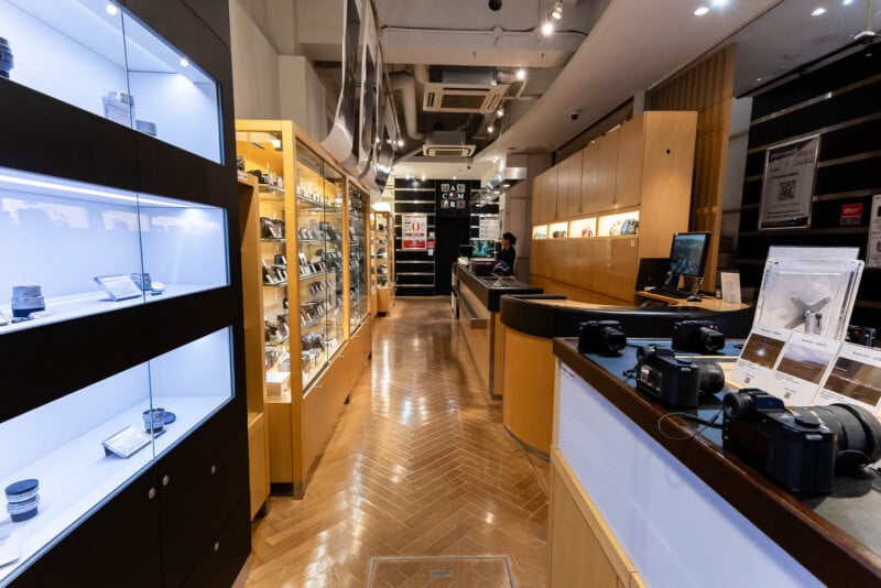 A modern camera store with glass display cases on both sides showing lenses and cameras. Wooden floors and bright lighting create a clean, organized atmosphere. A person stands behind a counter in the background.