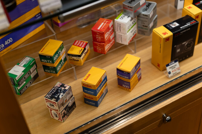Several boxes of 35mm film rolls, including brands like Kodak and Ilford, are displayed on a wooden shelf behind glass in a camera store. The boxes are various colors and stacked in rows.