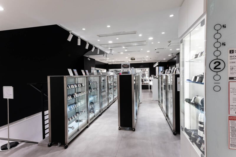A brightly lit electronics store displays rows of glass cases filled with cameras, lenses, and accessories. White walls and modern fixtures create a clean, organized shopping environment.