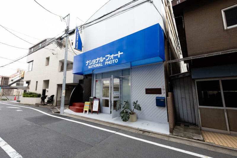 A small photo shop with a blue awning reading "NATIONAL PHOTO" in English and Japanese, situated on a quiet street corner between neighboring buildings in Japan. A bicycle is parked nearby.