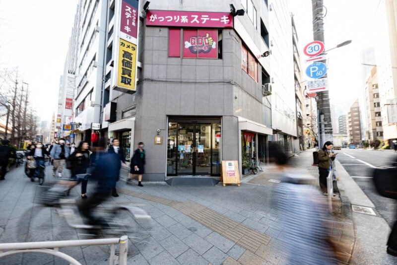 A busy street corner in a city with people walking and biking past a building that has Japanese signs, including a steakhouse sign above the entrance. The scene is lively with motion blur from pedestrians.