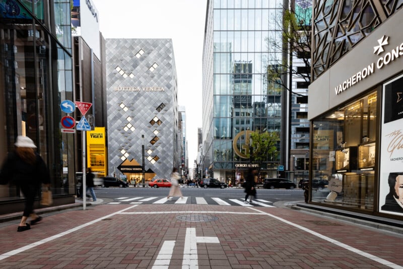 A city street intersection with pedestrians walking, luxury brand stores like Bottega Veneta, Giorgio Armani, and Vacheron Constantin, and tall glass buildings in an upscale shopping district.