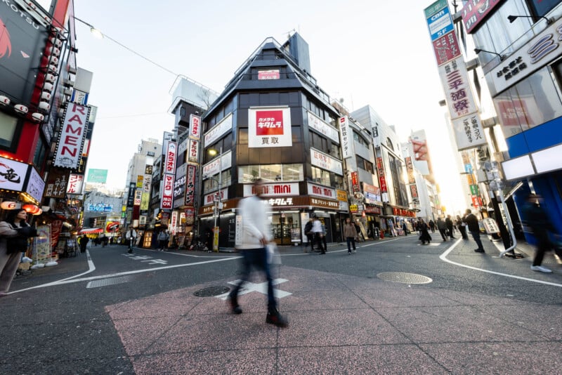 A busy city street in Japan with people walking, tall buildings covered in colorful signs, and bright advertisements in Japanese. The image captures an urban atmosphere at dusk with blurred motion of pedestrians.
