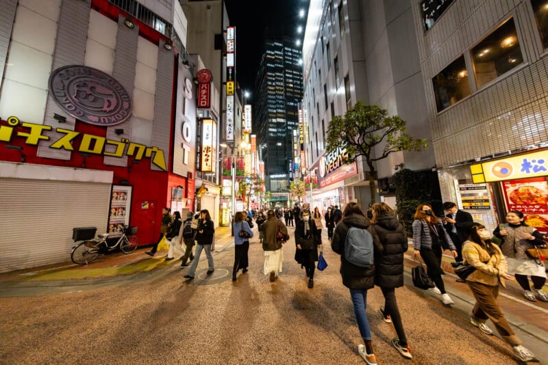 A busy city street at night with many people walking among bright neon signs and illuminated buildings in an urban area, likely in Japan.