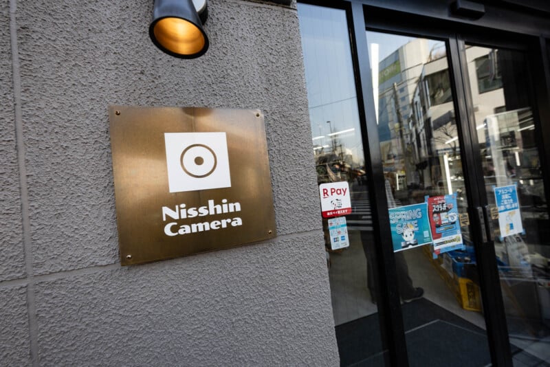 A brass sign reading "Nisshin Camera" is mounted on a textured gray wall next to a glass door with shop posters and payment stickers, reflecting buildings and trees outside.