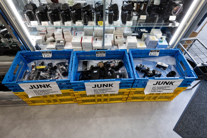 Three blue crates labeled "JUNK" filled with used camera equipment sit on yellow crates in a store. Shelves above display boxed items and cameras. Signs indicate no guarantee and cash only for items in the crates.
