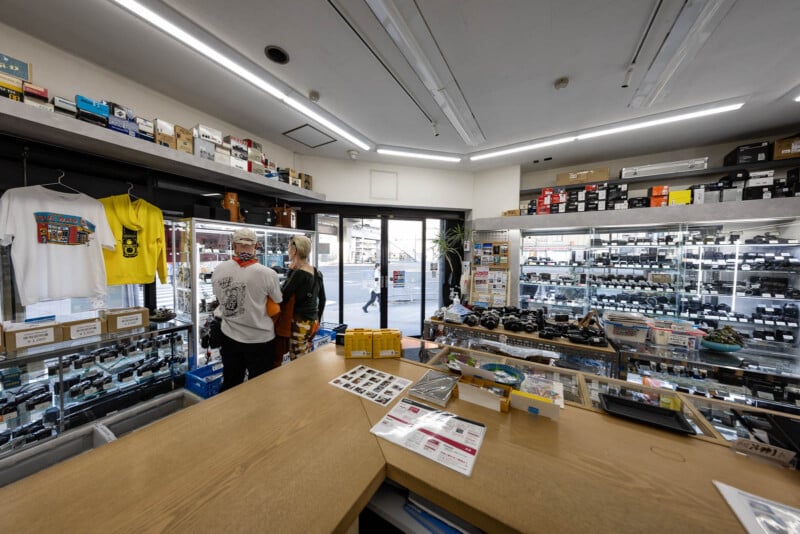 Interior of a camera shop with glass display cases filled with cameras and equipment, shelves lined with camera boxes, two people standing near the entrance, and a street visible outside through the glass door.