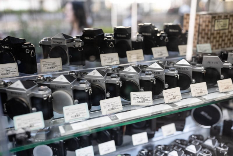 A glass display case shows several vintage Nikon film cameras arranged in rows, each labeled with price tags. The reflection of a blurred person is visible in the background.
