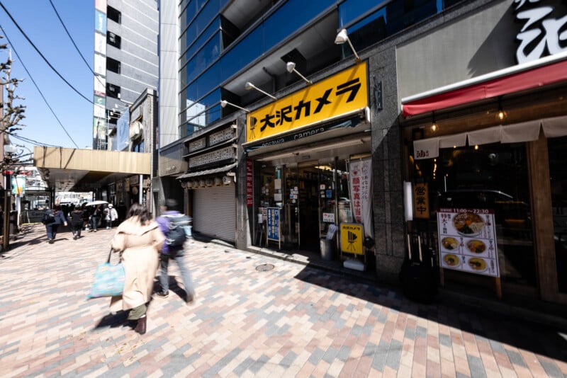 A city street in Japan with pedestrians walking past storefronts, including a camera shop with a yellow sign and a restaurant displaying a menu outside. Tall buildings and shops line the sidewalk on a sunny day.