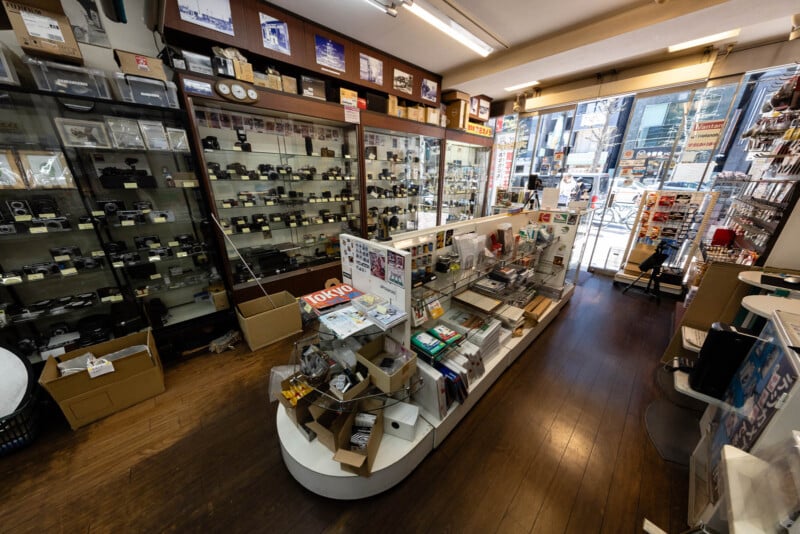 A camera shop interior with glass display cases filled with cameras and accessories, shelves of camera gear, magazines, and boxes. Bright natural light comes through the front windows facing a busy street outside.