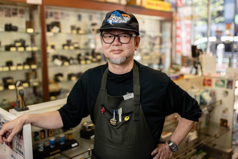 A man wearing glasses, a black cap, and a dark apron stands inside a camera shop, surrounded by display cases filled with cameras and photography equipment. He looks confidently at the camera with one hand on his hip.