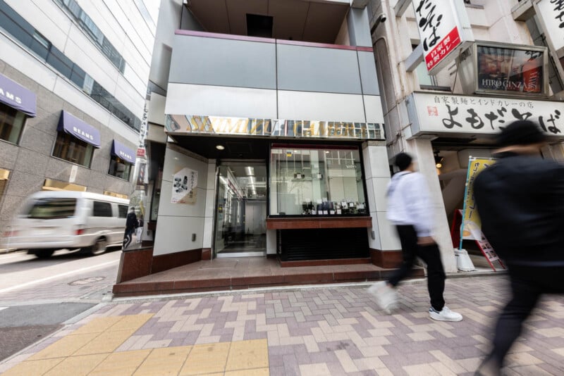 A street view of a modern storefront with glass display windows, showing watches inside. Two people walk past on the sidewalk, and nearby buildings with signs and awnings are visible.