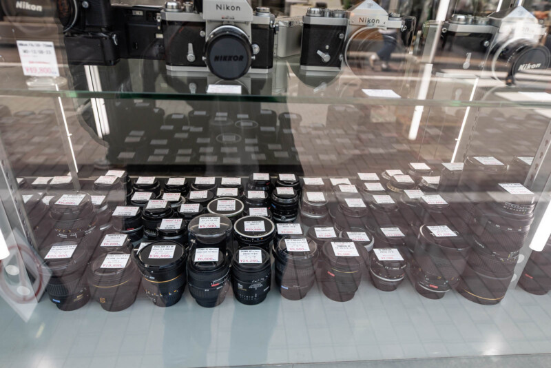 Glass display case with multiple camera lenses arranged in rows, price tags on top of each lens, and Nikon camera bodies on the shelf above. The scene is in a camera store with a street reflection visible in the glass.