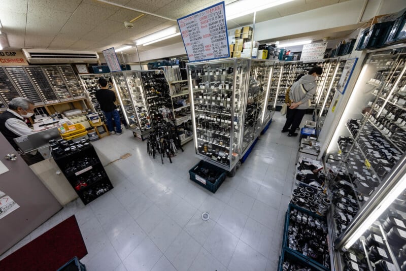 A shop with glass display cases filled with cameras and camera lenses; several people browse the merchandise under bright ceiling lights. The space is organized and well-lit.