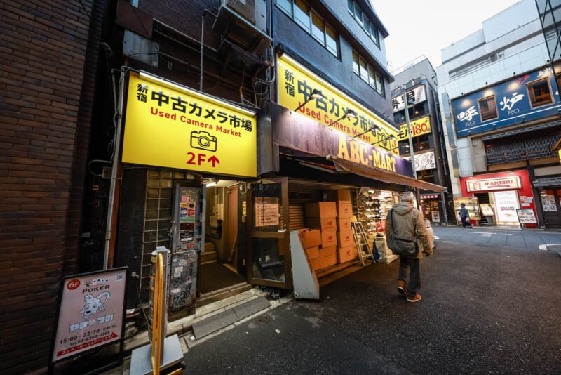 A person walks past a bright yellow sign for a used camera market in a narrow urban street lined with shops and signs in Japanese, with a stairway leading to the store’s entrance.