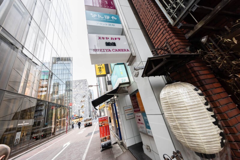A city street in Japan with modern glass buildings and a row of business signs, including one for "SUKIYA CAMERA." A large white paper lantern hangs near the brick wall on the right. The street is quiet and mostly empty.