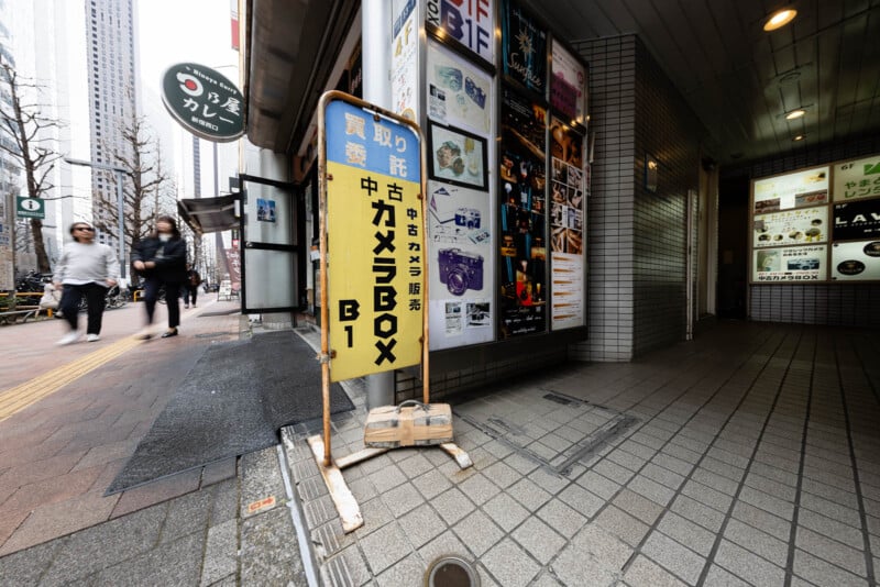 A yellow sign with Japanese text stands on a city sidewalk near a building entrance. Posters cover the wall behind it. People walk along the street, and tall buildings are visible in the background.