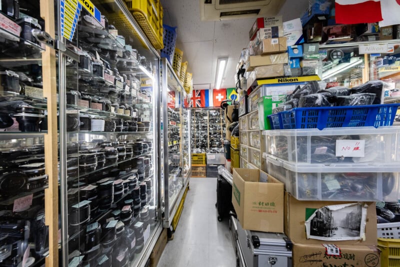 A narrow aisle in a cluttered camera shop, with glass display cases full of camera lenses on the left and shelves, boxes, and baskets of photo equipment on the right; various flags hang above the aisle.