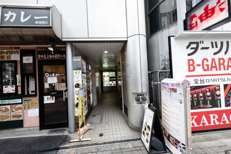 A narrow entrance to a building in Japan, with various restaurant signs, menus, and posters in Japanese on both sides. The hallway leads inside, and there are bright signs and displays on the walls.