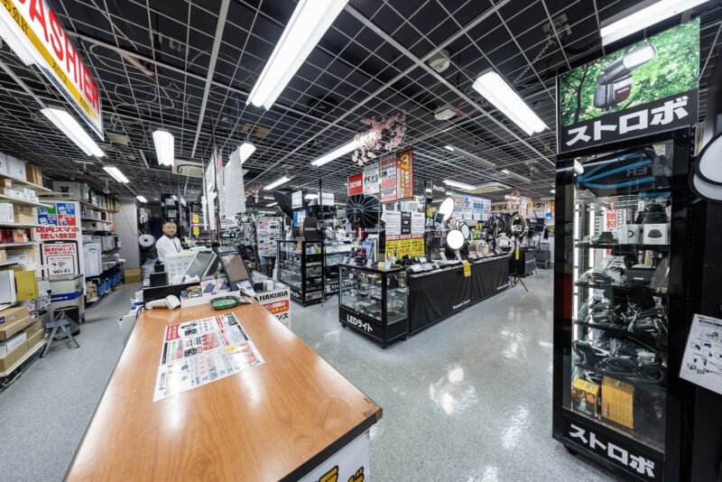 A well-lit camera store interior with display cases of photography equipment, cameras, and accessories. A person stands at the checkout counter on the left, while shelves and products fill the spacious, organized shop.